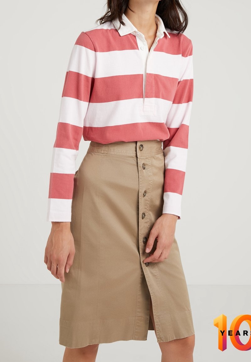 Gestreepte lange mouw polo in koraal en wit, gecombineerd met een beige rok met knopen aan de voorkant. De rok heeft een zijsplit en een gladde textuur.