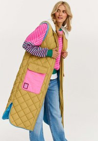 Prešívaný dlhý vest v olivovej zelenej farbe s ružovým vreckom a modrou podšívkou. Obsahuje kapucňu, veľké predné vrecko a voľný strih. Viditeľný detail pruhovaných rukávov.