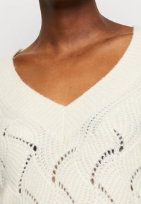 Pull en tricot de couleur crème avec un col en V, présentant un motif texturé et de subtils accents noirs. Matière douce et légère.