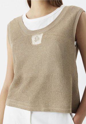 Beige strikket ærmeløs vest med tekstureret mønster, båret over en hvid t-shirt, med en lille udskæring med broderede detaljer ved halsudskæringen.