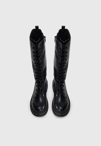 Bottes noires en cuir hauteur genou avec un devant à lacets, bout rond et semelle en caoutchouc. Doublure intérieure, dotées de languettes pour un enfilage facile.