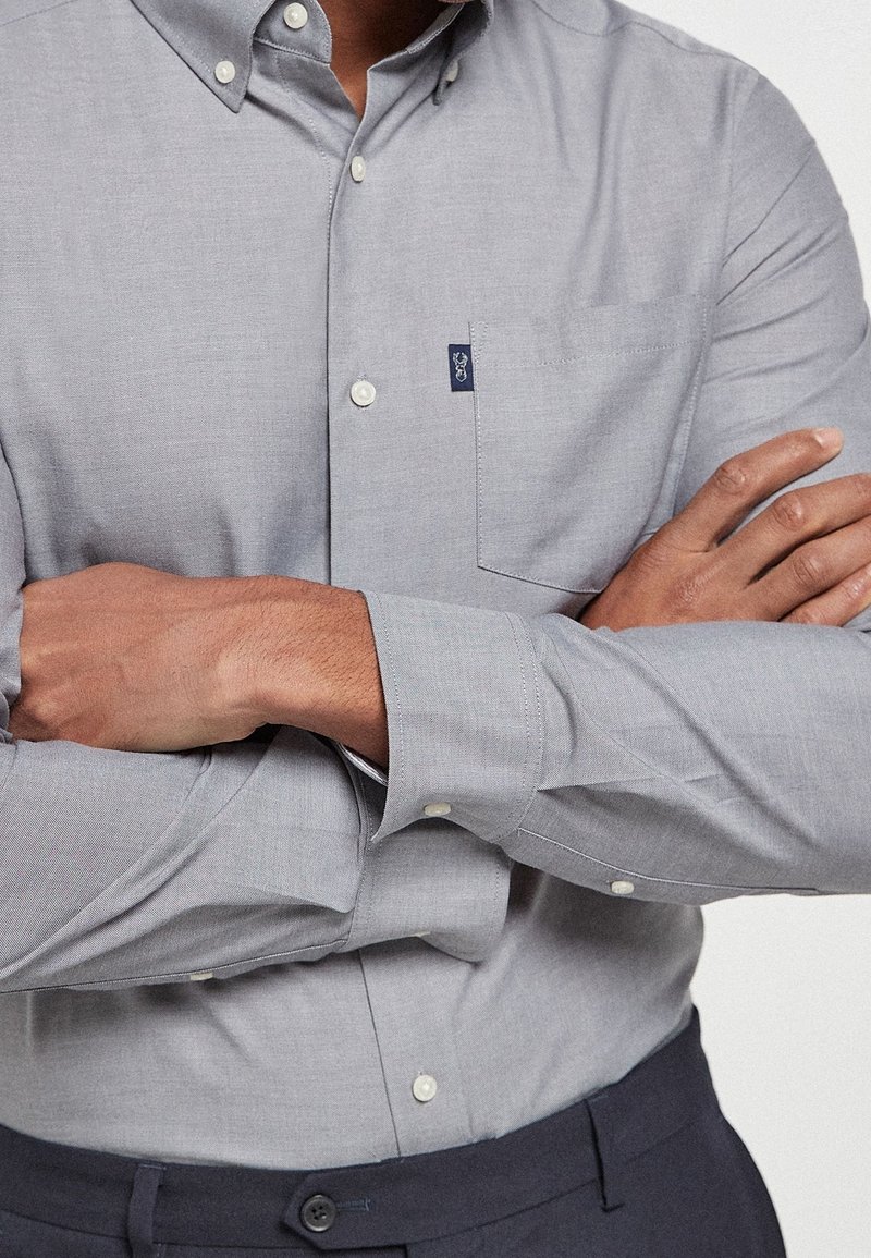 Homme portant une chemise gris clair boutonnée avec les manches longues croisées sur la poitrine, associée à un pantalon foncé.
