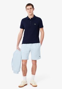 Polo nautique bleu marine avec logo vert, shorts bleu clair et veste bleu clair. Le modèle porte des baskets crème avec des accents marron.