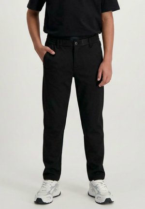 Persoon draagt zwarte broek en zwart shirt, met witte en grijze sneakers, staand met één hand in de zak tegen een effen achtergrond.