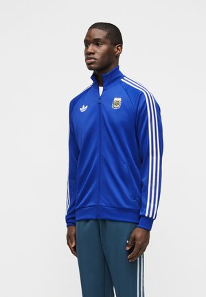 Fiatal férfi kék Adidas szabadidőfelsőt visel, fehér csíkokkal és az Argentína Labdarúgó Szövetség logójával, egyszínű háttér előtt áll.