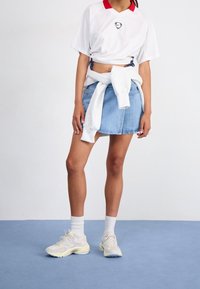 T-shirt blanc court avec col rouge et logo, associé à une mini-jupe en denim. Baskets de couleur claire avec accents texturés et chaussettes blanches.