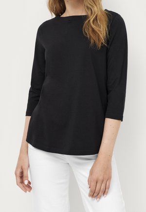 Femme portant un haut noir à manches trois-quarts associé à un pantalon blanc, debout devant un fond clair uni.