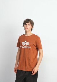 Smeđa pamučna majica s okruglim izrezom, koja ima bijeli logo s natpisom "ALPHA INDUSTRIES" i tri horizontalne pruge na prednjoj strani.
