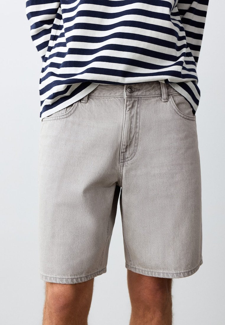 Personne portant un short en jean gris clair et une chemise à manches longues rayée bleu marine et blanc, mains dans le dos.