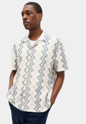 Junger Mann, der ein cremefarbenes kurzärmeliges Hemd mit blauen Zickzackmustern und dunkle Hosen trägt, steht mit den Händen in den Taschen und schaut seitlich.