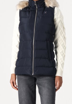 Vrouw draagt een marineblauw gestept vest met bontkraag over een witte kabelgebreide trui en donkergrijze jeans.