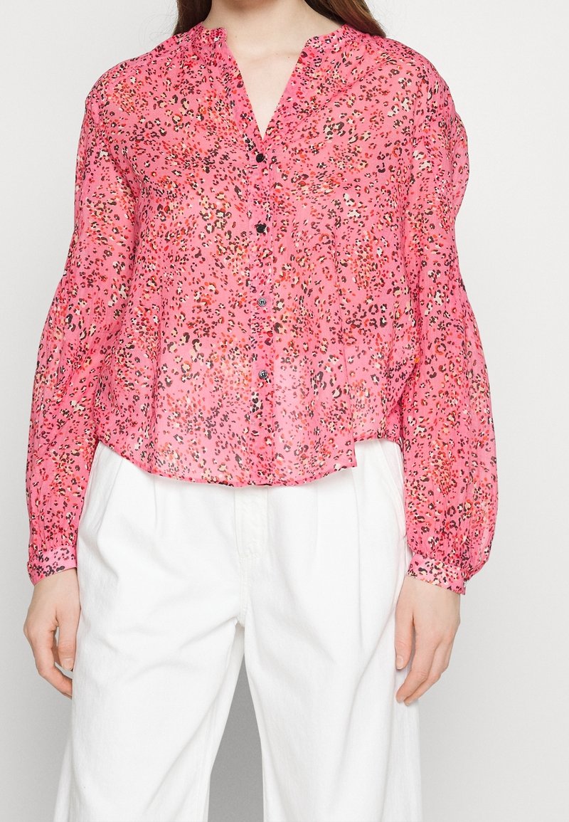 Femme portant un chemisier rose à imprimé léopard avec des manches longues bouffantes et un pantalon blanc taille haute.