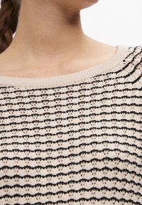 Close-up van de nek en schouders van een persoon die een beige gebreide trui draagt met een zwart horizontaal gestreept patroon en een ronde halslijn.