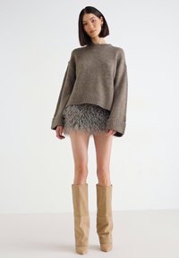 Overdimensjonert, olivenfarget strikkegenser med ribbestrikkede mansjetter, kombinert med et grått faux fur miniskjørt og beige høye hæler.