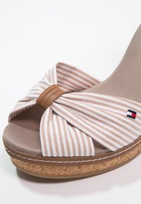 Sandale compensée à bout ouvert avec un nœud en tissu beige rayé blanc, semelle en liège et petit logo brodé rouge, blanc et bleu sur la bride.
