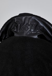 Chaqueta de shearling negra con una textura suave; cuenta con un interior de cuero negro con el logo "SIKSILK" grabado.