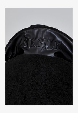 Melna flīsa jaka ar spīdīgu melnu ādas apkakli. Iespiests "SIKSILK" uzraksts. Mīksta tekstūra, minimālisma dizains.