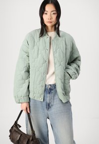 Mintgrønn quilted jakke med trykknapper, ribbede mansjetter og en avslappet passform, kombinert med lyseblå vide jeans og en mørkebrun håndveske.