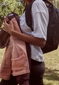 Black backpack on shoulder, holding a zip-up jacket in pink and maroon with mesh details, worn over a white t-shirt, and black leggings.