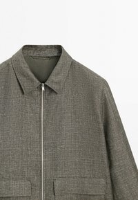 Grå lynlåstrøje med spids krave og to frontlommer med klap, vist på hvid baggrund.