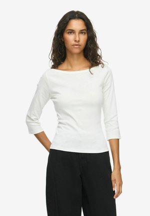 Femme aux cheveux bouclés et foncés, portant un haut blanc à manches trois-quarts et un pantalon noir, debout avec une main dans le dos.