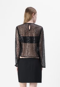 Schwarzes Pailletten-Oberteil mit langen Ärmeln, transparenter Textur und einem Schlüssellochdetail am Rücken. Kombiniert mit einem schwarzen Rock.
