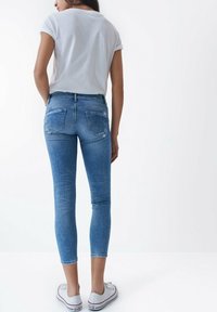 Jeans de mezclilla azul con un diseño ajustado, cintura de altura media, bolsillos traseros y sutiles detalles desgastados. Combinados con zapatillas blancas.