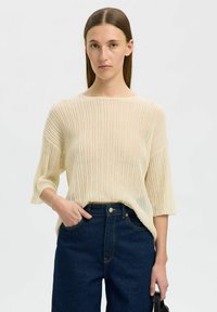 Cremfarbener, gerippter Strickpullover mit weiten Ärmeln, getragen über dunkelpulverblauen, hochgeschnittenen Jeans. Die Stricktextur ist weich und leicht.