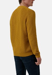 Musztardowy żółty sweter dzianinowy z teksturowanym wzorem, ze ściągaczami przy mankietach i hemie, przedstawiony od tyłu, zestawiony z ciemnoniebieskimi dżinsami.