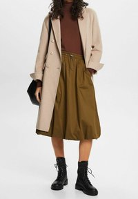 Manteau long beige sur un col roulé marron, associé à une jupe vert olive et des bottes noires à lacets. La jupe possède une taille élastique.