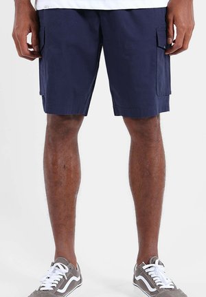 Homme portant un short cargo bleu marine et des baskets grises avec des lacets blancs, debout devant un fond blanc uni.