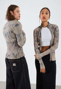 Ribbet zip-jakke i beige med blå tie-dye aksenter, kombinert med en hvit crop top og svart tettsittende skjørt. Modellene står rygg mot rygg.