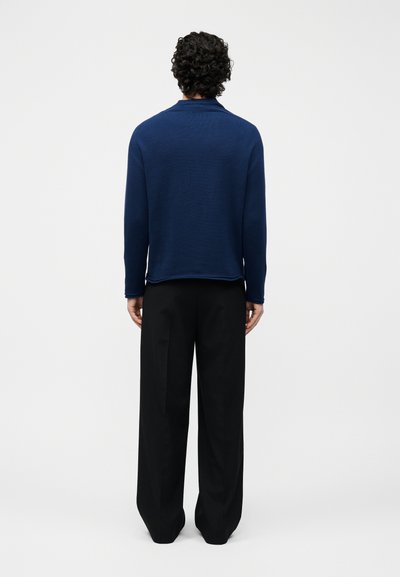 Άνδρας με σγουρά μαλλιά που φοράει μπλε μπλούζα μακρυμάνικη και χαλαρές μαύρες παντελόνες, στέκεται γυρισμένος προς τα πίσω σε λευκό φόντο.