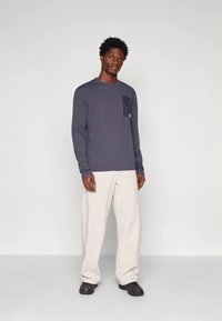 Mann in einem dunkelgrauen Langarmshirt mit Brusttasche, hellbeigen weit geschnittenen Hosen und schwarzen Schuhen, der vor einem weißen Hintergrund steht.