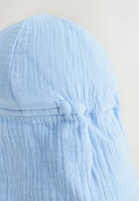 Svetlo modra kapa iz tkanine z teksturiranim vzorcem, zaobljenim vrhom in zbranimi detajli na zadnji strani z majhnim vozličkom.