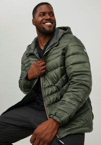 Chaqueta acolchada verde con capucha, que presenta un cierre con cremallera, bolsillos laterales y un acabado brillante. Camiseta negra visible por debajo.