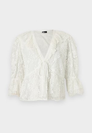 Bluza albă din dantelă cu mâneci bufante 3/4, decolteu în V, tiv cu volănașe și închidere cu snur în față.