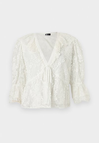 Blouse blanche en dentelle avec manches bouffantes 3/4, encolure en V, bordure à volants et fermeture avant à nouer.