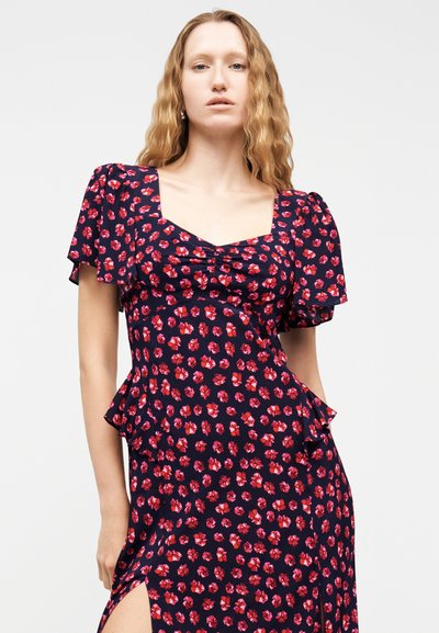 Γυναίκα με κυματιστά ξανθά μαλλιά φοράει μπλε σκούρο φόρεμα με κόκκινα ανθισμένα σχέδια, κοντά φουσκωτά μανίκια, τετράγωνη λαιμόκοψη και πλαϊνή σκίσιμο.