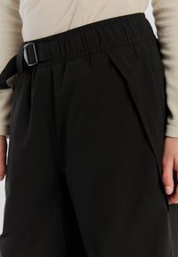 Zwarte, elastische broek met tailleband en riem, met een gladde textuur, een zijzak en een subtiele logo-detail op de linker bovenbeen.