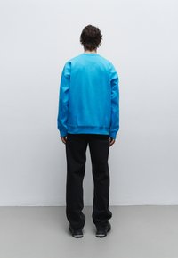 Blauwe oversized sweatshirt met een ronde halslijn en geribbelde manchetten, gedragen met zwarte broek en stevige zwarte schoenen, gezien vanaf de achterkant.