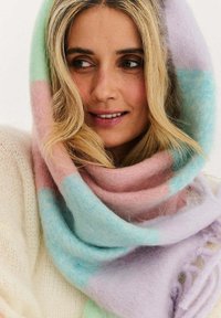 Blondynka z pastelowym, miękkim dzianym szalikiem owiniętym wokół głowy i szyi, patrząca w bok z lekkim uśmiechem.