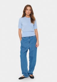 Light blue knitted short-sleeve sweater with a textured pattern. Loose-fitting blue denim pants with a relaxed silhouette and pockets. Black flat shoes.