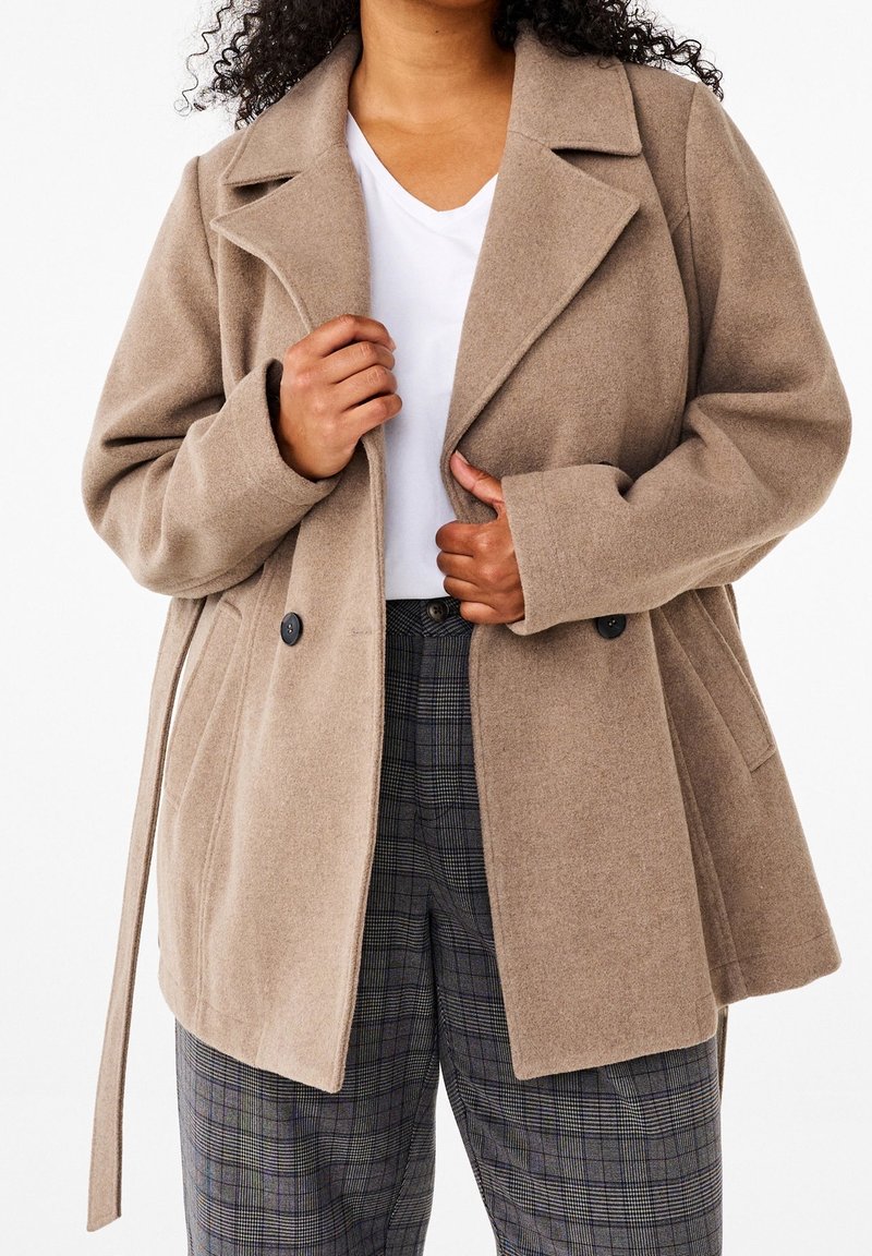Mujer con un abrigo de lana beige sobre una camiseta blanca y pantalones de cuadros grises, ajustándose el abrigo por delante con ambas manos.