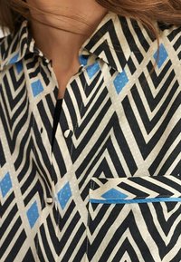 Camisa de botones con un patrón en zigzag en negro, blanco y azul; cuenta con un cuello puntiagudo y un bolsillo en el pecho con un acento azul.