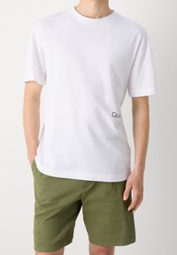 Biała bawełniana koszulka z okrągłym dekoltem, krótki rękaw i małe czarne logo z przodu; zestawiona z oliwkowymi szortami.