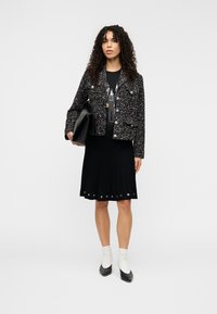 Veste en tweed noire à boutons métalliques portée sur un tee-shirt graphique noir, associée à une jupe plissée noire ornée d'œillets et des chaussures à talons.
