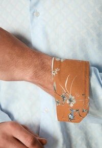 Mouw van een lichtblauw overhemd met knopen en een textuurpatroon, voorzien van een oranje bloemenprint als accent op de mouw met blauwe stikdetails.