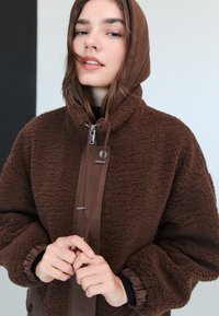 Ruskea fleecetakki, jossa korkea kaulus ja huppu. Siinä on sileät nahkakoristeet, vetoketju ja joustavat hihansuut. Pinta on teksturoitu ja materiaali lämmin.