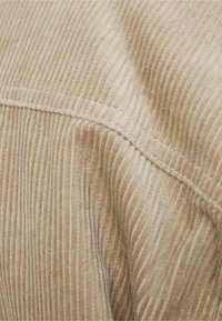 Béžová manšestrová tkanina s jemnou žebrovanou strukturou, viditelným šitím a záhybem, zdůrazňující její poddajný a teplý design.